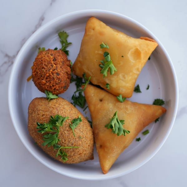 A white bowl holds two golden samosas, a falafel, and a kibbeh, garnished with parsley.