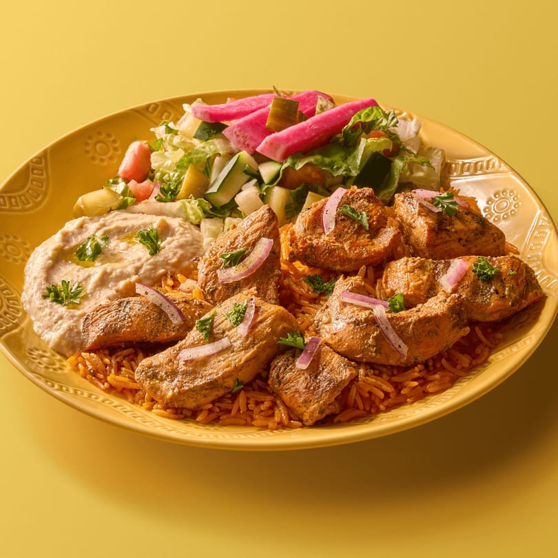 Grilled chicken and orange rice with hummus, fresh salad, and pink pickled turnips on a yellow plate.