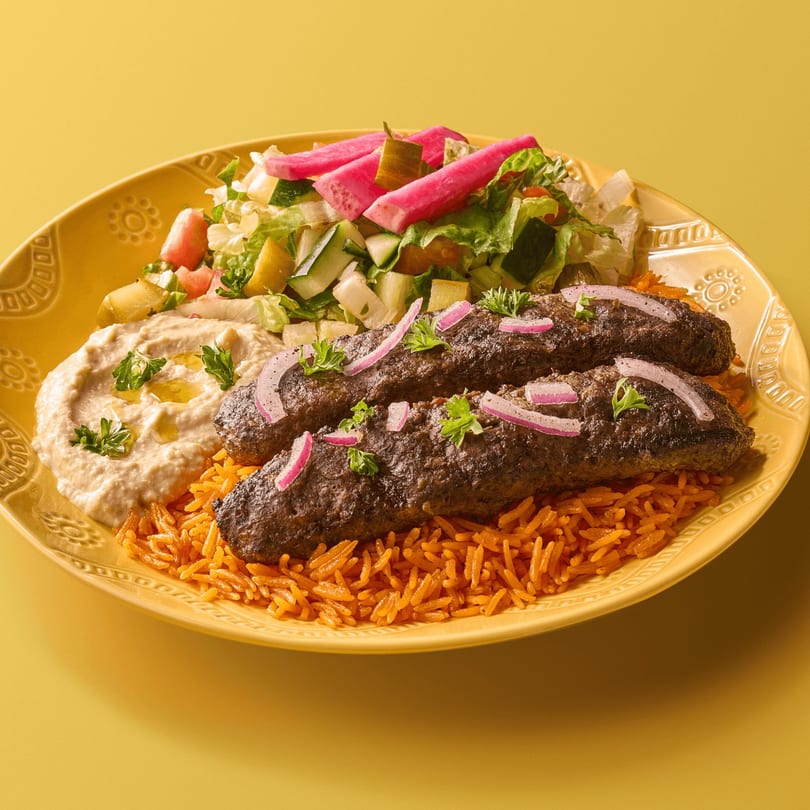 Two kebabs, orange rice, hummus, and a salad with pink pickled vegetables on a yellow plate.